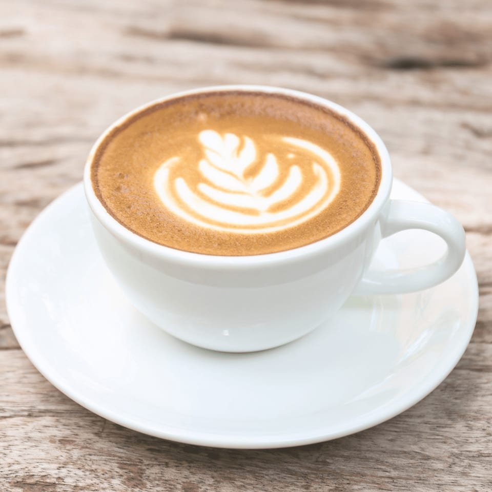 A white cup of latte with a leaf design in the foam, on a wooden table.