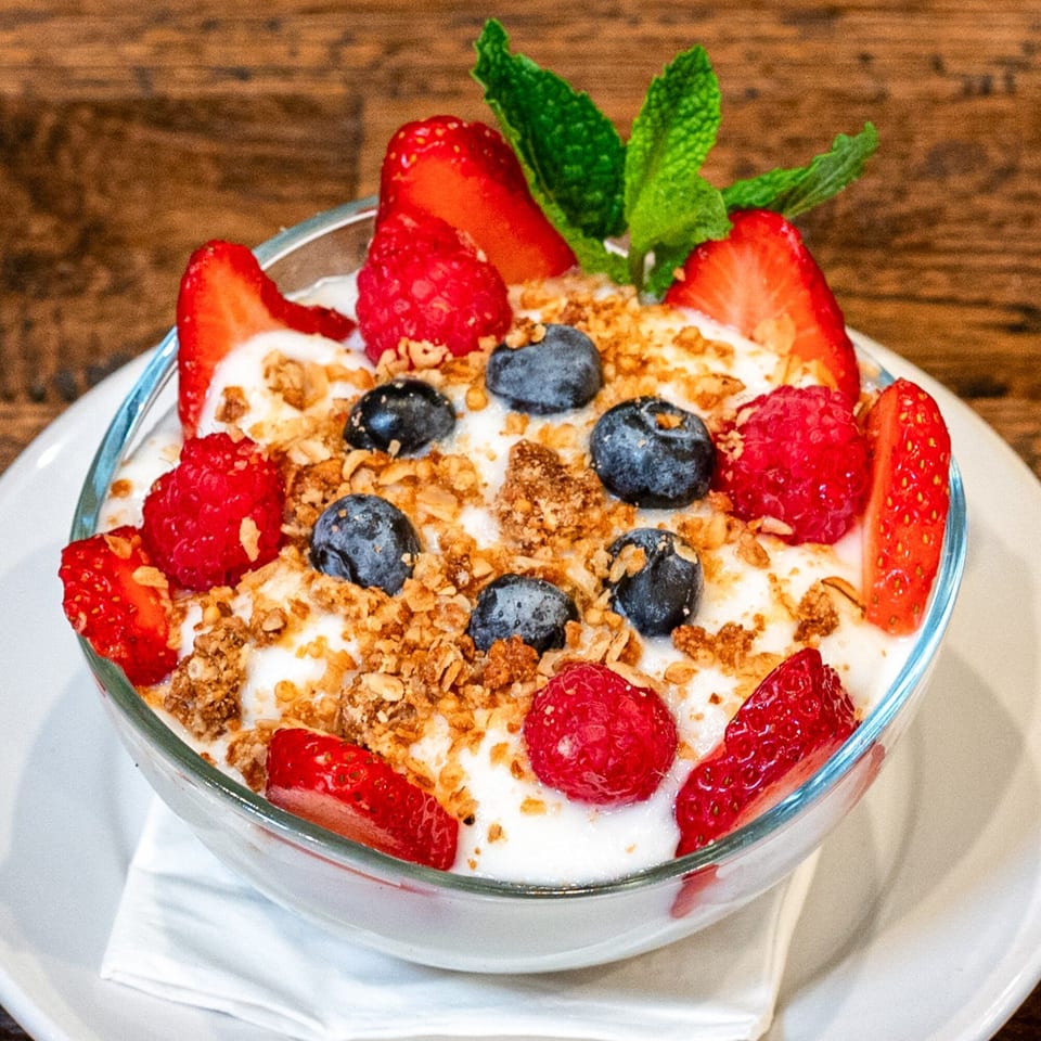 A glass bowl of yogurt parfait with strawberries, blueberries, raspberries, granola, and mint.