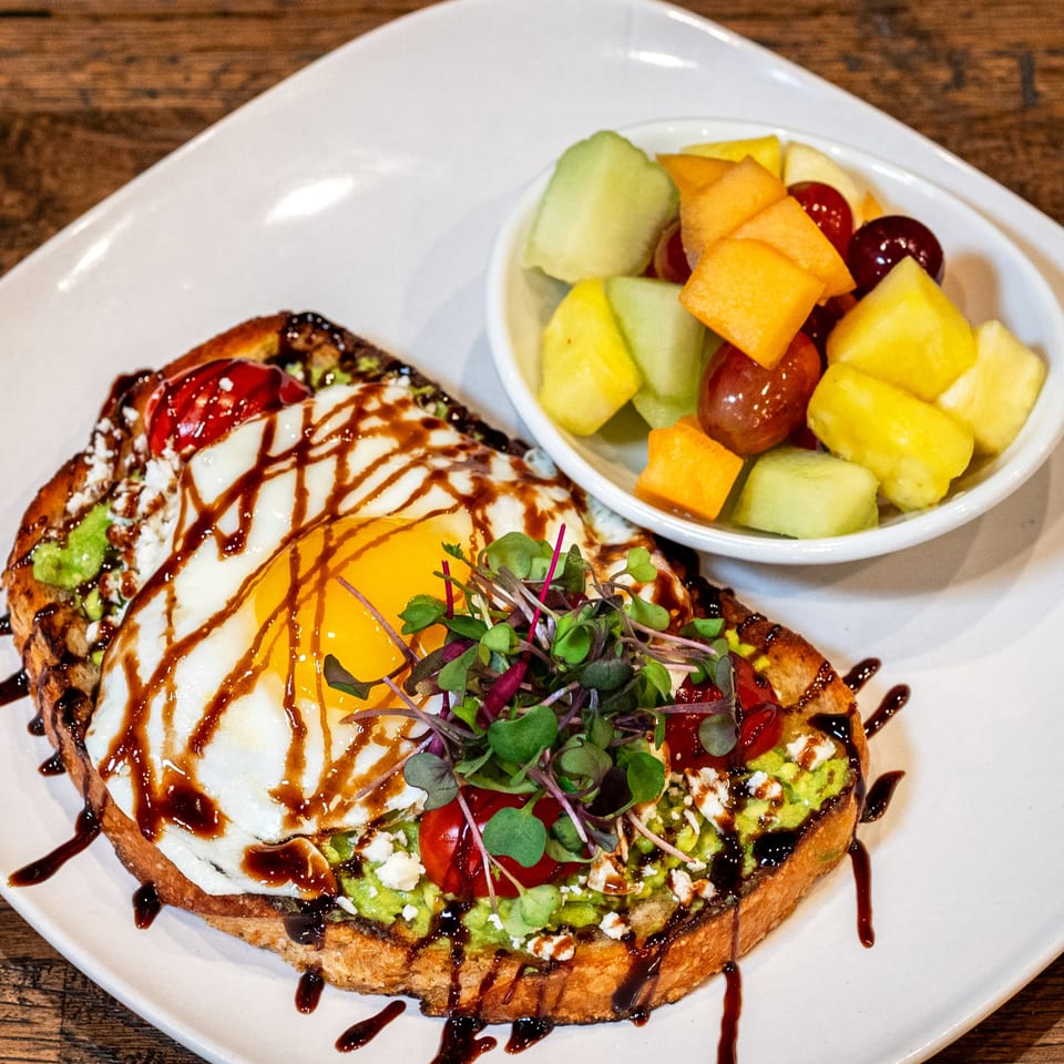 Avocado toast with a fried egg, cherry tomatoes, feta, balsamic glaze, and a side of fruit.