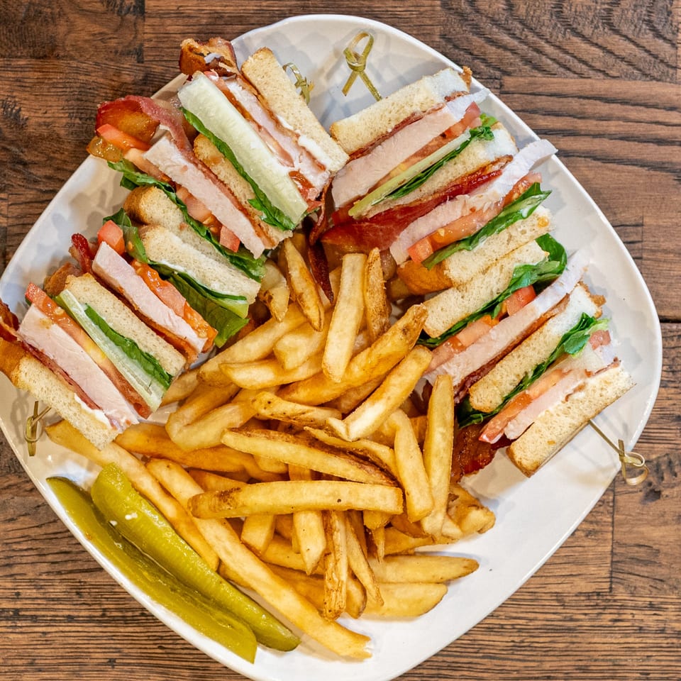 A Brunchy's Cafe club sandwich, cut into quarters, served with golden fries and pickle spears.