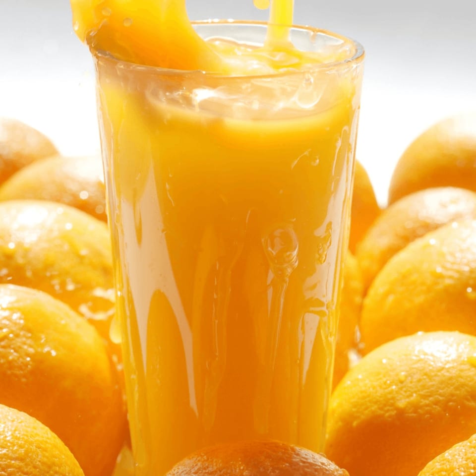 Fresh orange juice being poured into a glass, surrounded by whole oranges.