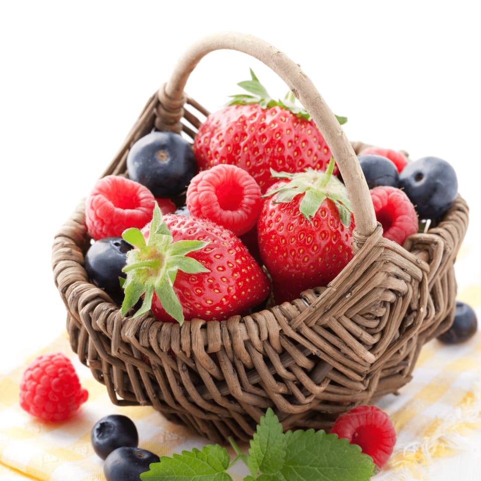 A wicker basket overflowing with fresh strawberries, raspberries, and blueberries on a checkered cloth.