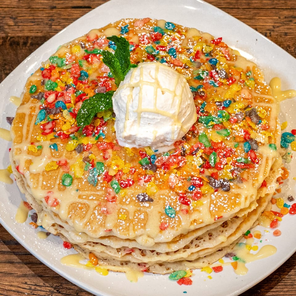 Stack of pancakes topped with colorful cereal, white icing, powdered sugar, whipped cream, and mint.
